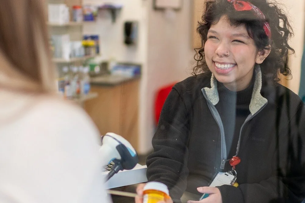 Un miembro del personal de la farmacia « Mosaic », con una sonrisa, atiende a un paciente en el mostrador de la farmacia