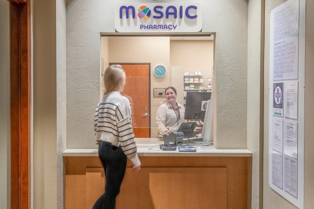 Empleado de farmacia sonriendo mientras atiende a un paciente en la ventanilla de atención al público Mosaic , con los estantes de medicamentos visibles detrás del mostrador.