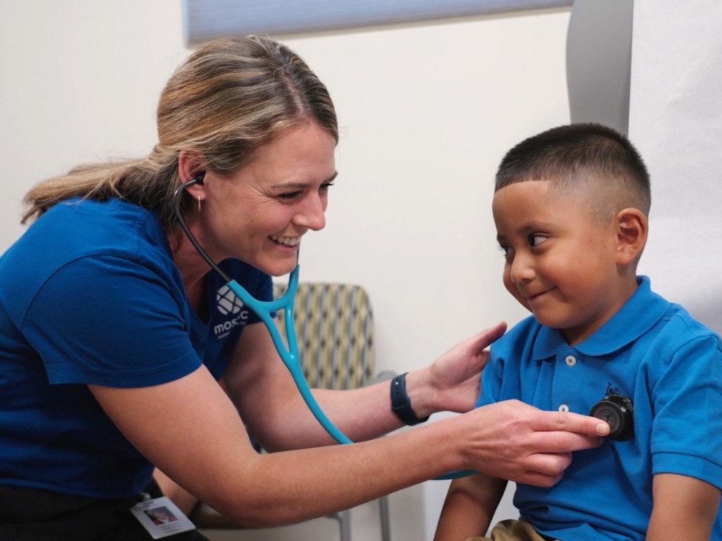 A Mosaic healthcare provider uses a stethoscope to examine a smiling young child during a pediatric visit.