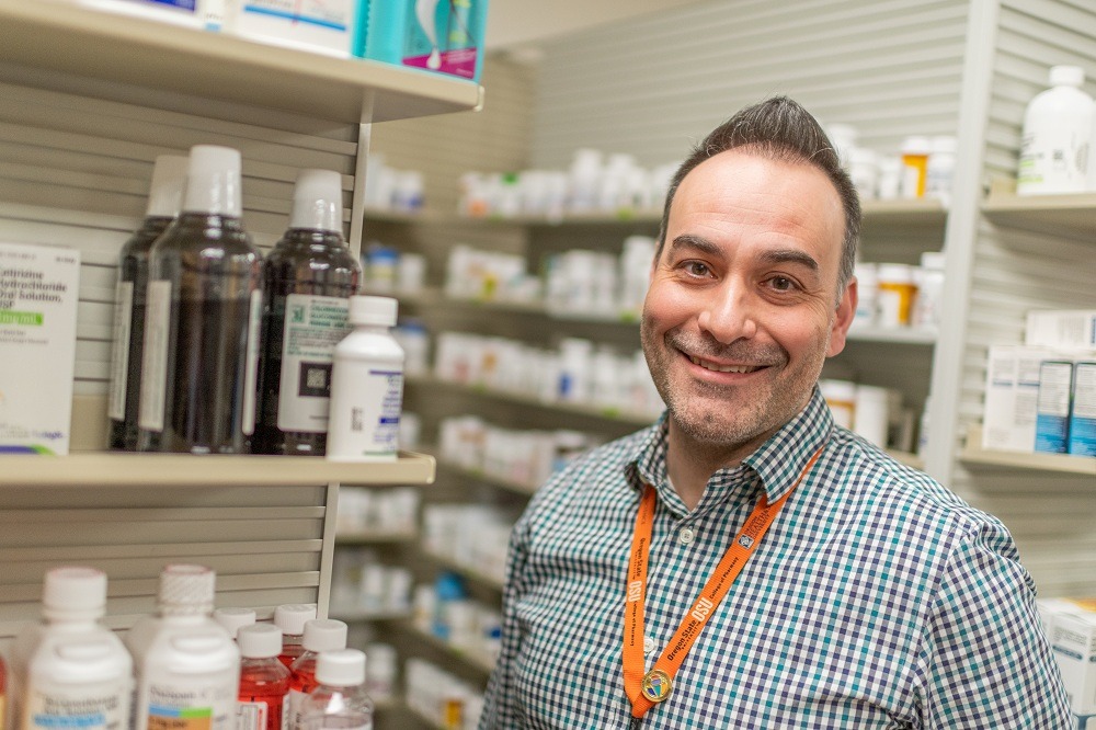 Miembro del equipo de farmacia sonriendo mientras está de pie frente a los estantes de medicamentos dentro de una farmacia minorista, con frascos de medicamentos recetados visibles al fondo.