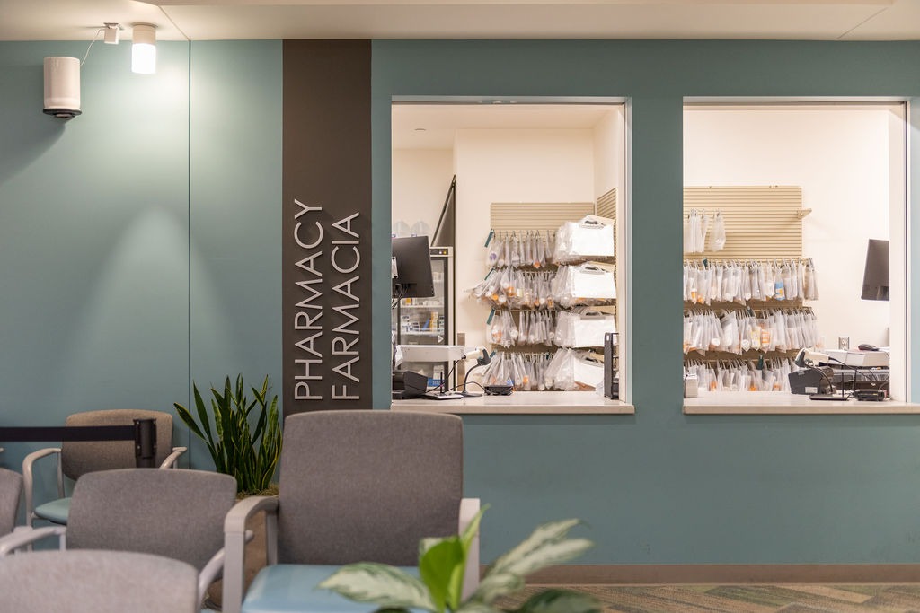Sala de espera con sillas frente a dos ventanillas de servicio de farmacia en un centro Mosaic Center, con un letrero que dice «Pharmacy / Farmacia» en la pared y bolsas con medicamentos recetados visibles detrás del mostrador.