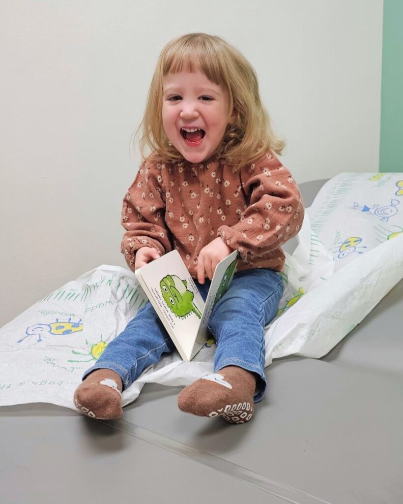 Smiling young child sitting on an exam table in a clinic, holding and looking at a small board book during a pediatric visit.