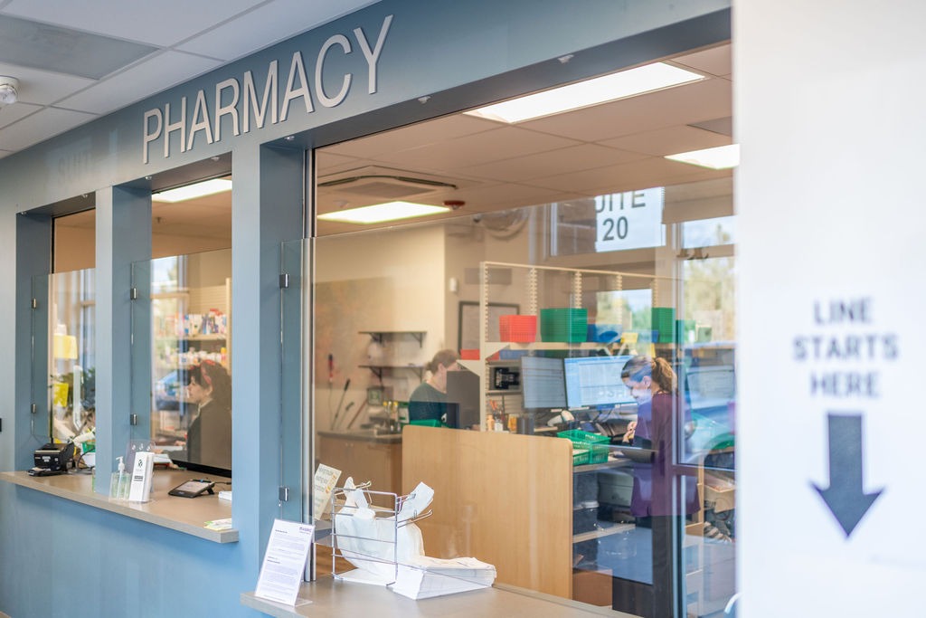 Empleados de farmacia trabajando detrás del mostrador en una Mosaic , con una ventanilla de atención al público y el letrero «Farmacia» visible sobre el mostrador.