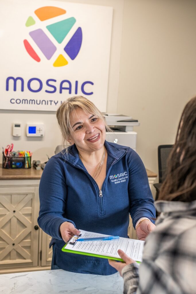 A Mosaic Community Health staff member smiles while handing a clipboard with paperwork to a patient at a front desk, with the Mosaic logo displayed on the wall behind her.