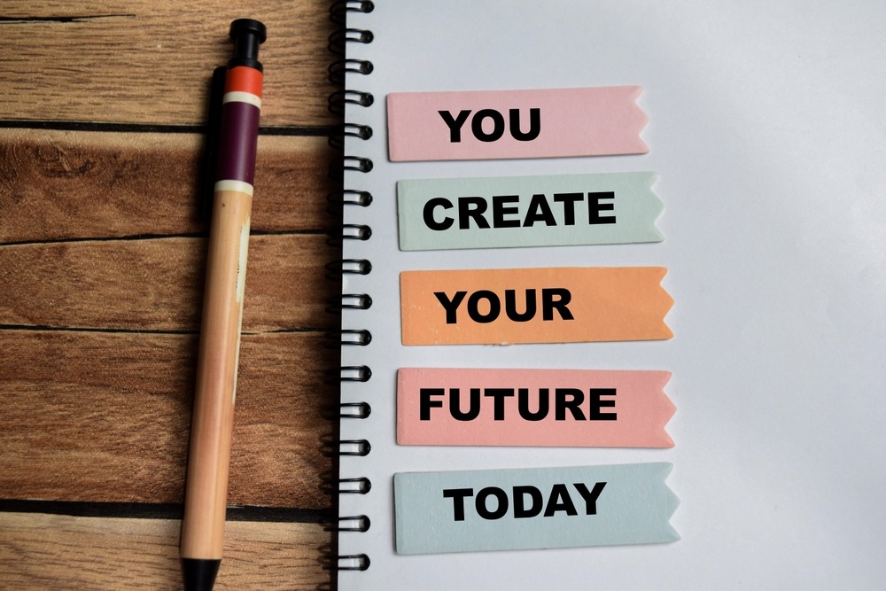 Notebook with tabs reading ‘You create your future today’ next to a pen on a wooden desk.